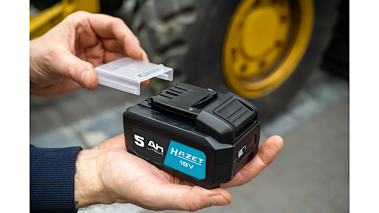 Zwei H&auml;nde halten einen schwarzen 18V-Akku mit blauer Beschriftung, w&auml;hrend ein transparenter Schutzdeckel angebracht oder entfernt wird. Hintergrund unscharf mit Werkstattumgebung.